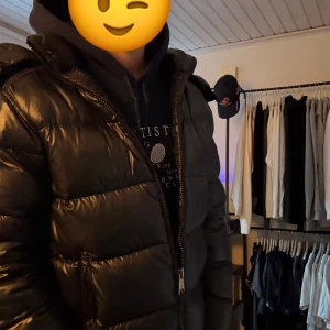 Svart Ralph Lauren pufferjacka  - Säljer en svart pufferjacka från Ralph Lauren med huva och dragkedja framtill. Jackan har en glansig yta, breda quiltade sektioner och två sidofickor med dragkedja. Perfekt för kalla dagar och har en clean, modern look. Köpt på NK för ca 7500