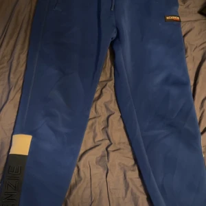 Blå mjukisbyxor från McKenzie - Snygga blå mjukisbyxor från McKenzie med svarta detaljer och stort broderat McKenzie-tryck längs benen. Byxorna har elastisk midja och är perfekta för chill eller träning. Materialet är mjukt och bekvämt, och designen är modern med en sportig vibe. Kom med bud