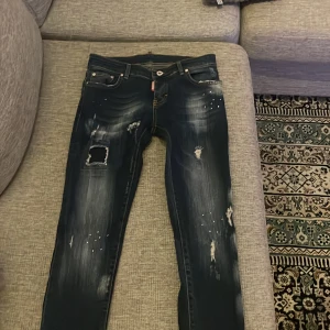 Dsquared2 blå slitna jeans med detaljer - Säljer ett par blå jeans från Dsquared2 med coola slitningar, patch med italienska flaggan bak och små nitar på benen. Jeansen har skinny passform och klassisk femficksdesign. Perfekta för dig som gillar streetstyle och unika detaljer.
