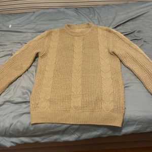 Beige stickad tröja med kabelmönster - Säljer en beige stickad tröja med klassiskt kabelstickat mönster framtill. Tröjan har rund halsringning och långa ärmar, perfekt för kyliga dagar. Den är mjuk och skön, passar till både jeans och kjol för en avslappnad look. 