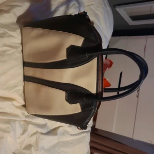 Svart och beige handväska i skinnimitation - Snygg handväska i svart och beige med stilren design och rejäla handtag. Väskan har dragkedja upptill och ett rymligt foder i satinliknande material. Perfekt för dig som vill ha en klassisk och trendig accessoar med plats för allt du behöver.