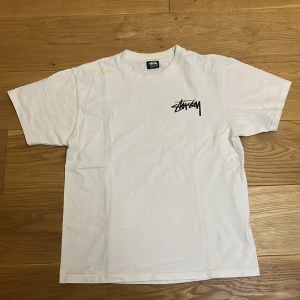 Vit Stüssy t-shirt med tärningsprint - Vit t-shirt från Stüssy i bomull med klassisk logga på bröstet och stor grafisk print med rosa tärningar och svart Stüssy-tryck på ryggen. Kortärmad och normal passform, perfekt för en streetwear-look.