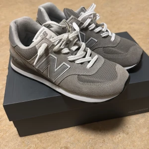 Grå New Balance 574 sneakers - Snygga New Balance 574 sneakers i grått med vita snören och klassisk N-logga på sidan. Ovandel i mocka och mesh för en sportig vibe och bra andningsförmåga. Skorna har svart yttersula med grovt mönster och rund tå. Perfekta för dig som gillar stilrena och bekväma sneakers. (Säljer dem för en sko har en lite mörkare färg men man ser inte mycket skriv om due intresserad)