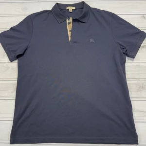 Mörkblå Burberry Brit pikétröja - Snygg mörkblå pikétröja från Burberry Brit i 100% bomull. Klassisk krage, korta ärmar och ikonisk rutdetalj vid knäppningen. Liten broderad logga på bröstet. Perfekt för en clean och stilren look.