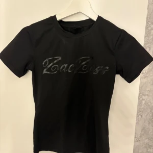 Svart ZacZess t-shirt med tryck - Svart t-shirt från ZacZess med glansigt svart logotyp-tryck framtill. Klassisk rund halsringning och korta ärmar. Tight passform och tillverkad i mjukt material som känns skönt mot huden. Perfekt för dig som gillar stilrena och enkla plagg med en twist.
