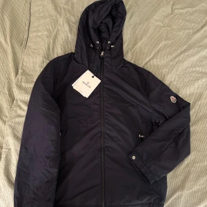 Mörkblå vindjacka från Moncler - Snygg mörkblå vindjacka från Moncler med huva och dragsko, hel dragkedja framtill och två fickor med dragkedja. Jackan har Moncler-logga på ärmen och är tillverkad i lätt polyester, perfekt för blåsiga och regniga dagar.