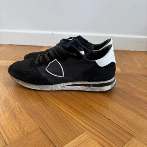 Svarta sneakers från Philippe Model - Svarta sneakers från Philippe Model med vita detaljer på hälen och sidorna. Skorna har snörning, rund tå och platt sula. Ovandelen är i en mix av mocka och textil, vilket ger en snygg kontrast. Perfekta för dig som gillar en stilren och sportig look.