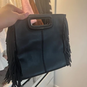 Svart handväska med fransar från Maje - Cool svart handväska från Maje i skinn med rektangulärt handtag och snygga fransar längs sidorna. Väskan har en stilren design med en vertikal söm framtill och är perfekt för att lyfta din outfit. Rymlig insida och smidigt handtag gör den enkel att bära med sig. Man får även med bandet och dustbag.