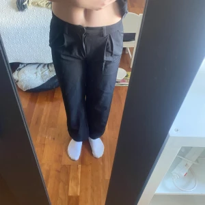 Svarta vida kostymbyxor - Säljer ett par svarta kostymbyxor med hög midja och vida ben. Byxorna har snygga veck framtill och stängs med dragkedja och knapp. Perfekta om du vill ha en clean och stilren look. Passar till både sneakers och boots. Storlek 146