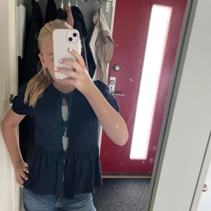 Marinblå blus med knyt och volang - Säljer en marinblå blus med korta puffärmar och volang nedtill. Blusen har tre snygga knyt framtill. Perfekt att styla med jeans eller kjol för en trendig look.