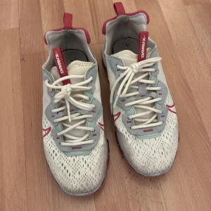 Nike React Element 87 D/MS/X sneakers - Nike React Element 87 D/MS/X sneakers i vitt mesh med ljusgrå och mintgröna detaljer, röda accenter och snörning. Snygg design med genomskinliga partier och röda swoosh-loggor på sidorna. Skön och futuristisk sula med orange detaljer. Perfekta för dig som gillar streetwear.