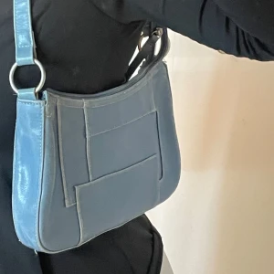 Blå axelväska i skinn med detaljer - Snygg blå axelväska i skinn med unika geometriska sömdetaljer på framsidan. Väskan har bred axelrem med silvriga ringar och dragkedja upptill. Perfekt storlek för att få plats med allt du behöver till vardags.