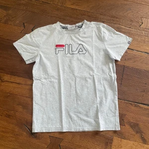 Grå FILA t-shirt med logga - Snygg grå t-shirt från FILA med klassisk logga i rött, blått och vitt på bröstet. Modellen är rak med rund hals och korta ärmar. Perfekt för dig som gillar sportiga och stilrena plagg. Mjuk och skön bomull som funkar året runt.