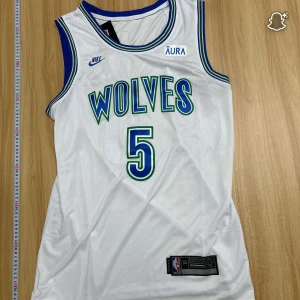 Vit Wolves baskettröja Nike Edwards #5 - Snygg vit baskettröja från Nike med blå och gröna detaljer, Wolves-tryck på framsidan och Edwards samt nummer 5 på ryggen. Ärmlös modell med blå/gröna ränder runt hals och ärmhål. Officiell NBA patch nedtill. Perfekt för dig som gillar basket och streetwear.