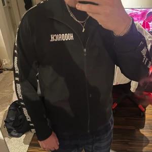 Svart ziptröja från Hoodrich - Svart ziptröja från Hoodrich med vit logga på bröstet och längs ärmarna. Tröjan har hel dragkedja framtill, ståkrage och långa ärmar. Perfekt för en streetwear-look och enkel att matcha med andra plagg.