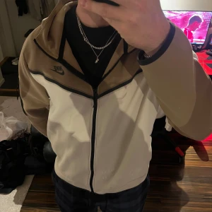 Nike tracksuit tröja i beige och vitt - Nike ziptröja med huva i beige och vitt, svart Nike-logga på bröstet och svarta detaljer. Tröjan har hel dragkedja framtill och en extra ficka med dragkedja på ärmen. Perfekt för dig som gillar sportig och clean stil. Storlek XL men passar mig som har M/L