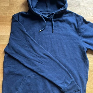 Marinblå hoodie från 157 – Storlek S - Stilren och bekväm hoodie i marinblått från 157. Tröjan har huva med justerbar snörning, praktisk magficka och ribbade muddar för en bra passform. Tillverkad i mjukt material som känns skönt mot huden – perfekt för vardagsbruk eller avslappnade dagar.  Skick: Mycket bra / knappt använd Storlek: S 