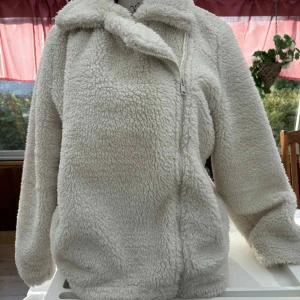 Vit teddyjacka från Threadbare - Mysig teddyjacka i vit färg från Threadbare, med fluffig yta och mjukt foder. Jackan har en sned dragkedja framtill, stor krage och långa ärmar. Perfekt för dig som gillar chill och comfy stil under kyliga dagar.