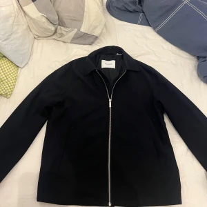 Overshirt från Les Deux med dragkedja - Stilren svart jacka från Les Deux i storlek M. Jackan har klassisk krage, hel dragkedja framtill och diskret broderad logga på bröstet. Köpt för 2300kr. Skick 10/10 inte kommit till användning. Overshirt perfekt för hösten. Mitt pris 900kr. Stilren passar till allt. Vid frågor eller intresse hör av dig!🫡