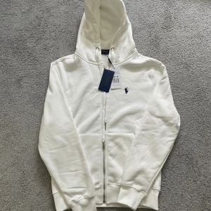 Vit zip/hoodie från Polo Ralph Lauren - Vit hoodie från Polo Ralph Lauren med dragkedja och snörning i huvan. Tröjan har ett mörkblått broderat logomärke på bröstet och är tillverkad i mjuk bomull med fleecefoder på insidan. Klassisk passform med långa ärmar och ribbade muddar. Den är i storlek M men passar storlek S.