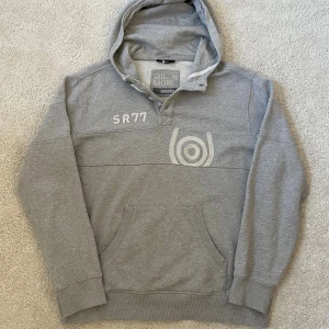 Grå hoodie Sail Racing | XL - Snygg grå hoodie från Sail Racing i storlek XL med broderad SR77-logga och tryck på bröstet. Tröjan har huva, knappar vid halsen och en stor magficka. Mjuk insida och ribbade muddar gör den extra skön. Perfekt för chill dagar.
