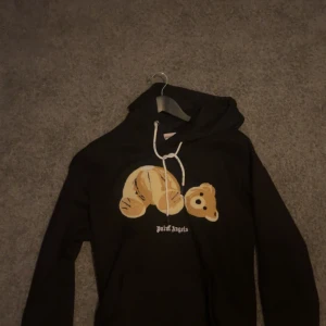 Svart Palm Angels hoodie  - Svart hoodie från Palm Angels med ikoniskt björnmotiv på bröstet och vit snörning i huvan. Tröjan har lång ärm och klassisk känguruficka framtill. Perfekt statement-plagg för dig som gillar streetwear och coola prints.
