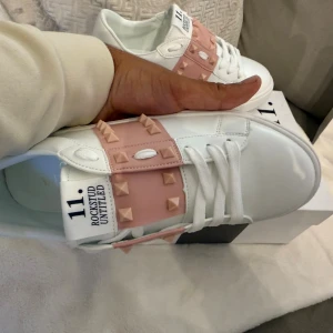 Valentino Rockstud sneakers vit/rosa - Snygga Valentino Rockstud sneakers i vitt skinn med bred rosa rem över foten dekorerad med ikoniska nitar. Klassisk rund tå, platt sula och vita snören. Märkeslogga på plösen och detaljerade nitar även på hälen. Perfekta för dig som vill sticka ut med lyxig streetstyle.