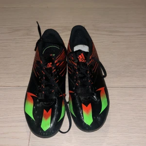 Adidas Messi barnfotbollsskor svart/röd - Säljer ett par Adidas Messi fotbollsskor i svart och rött med gröna detaljer. Skorna har snörning, mönstrad ovandel och dobbar i grönt för bra grepp på planen. Snygg design med Messi-logga och Adidas-tryck. Perfekta för dig som vill sticka ut på fotbollsplanen.
