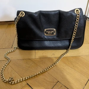 Svart axelväska Michael Kors skinn - Snygg svart axelväska från Michael Kors i mjukt skinn med guldfärgade kedjedetaljer och logoplatta framtill. Väskan kortfack samt andra flera fack och innerfoder med Michael Kors-tryck. Perfekt storlek för det viktigaste och stilren design. Köpt på Nordstrom i USA. 