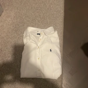 Vit skjorta från Ralph Lauren - Vit klassisk skjorta från Ralph Lauren med blå broderad logga på bröstet. Skjortan har knappar framtill och en stilren krage. Perfekt för dig som gillar enkel och tidlös stil. 