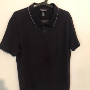 Svart pikétröja från H&M slim fit - Klassisk svart pikétröja från H&M i slim fit-modell. Tröjan har korta ärmar, krage med vit piping och knäppning framtill. Perfekt för en clean och stilren look. Materialet känns mjukt och bekvämt mot huden.