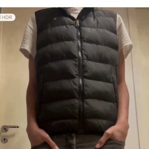 Svart dunväst från Gant - Svart dunväst från Gant med quiltad design och hög krage. Västen har två sidofickor och stängs med dragkedja framtill. Perfekt för lager-på-lager och har en stilren look med diskret logga på bröstet. Kom dm för frågor, pris kan diskuteras.
