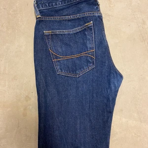 Mörkblå jeans från Hollister W32 L32 - Snygga mörkblå jeans från Hollister med klassisk femficksdesign och kontrastsömmar. Modellen har rak passform och normal midja, tillverkade i slitstarkt denim med detaljerade nitar och knappstängning. Perfekt för dig som gillar en enkel och tidlös stil.