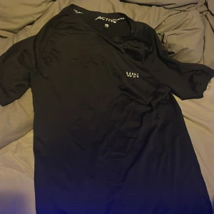 Svart MAN Active t-shirt i S - Svart t-shirt från MAN Active i storlek S. T-shirten har korta ärmar och en diskret logga på bröstet. Materialet är mjukt och lätt polyester, perfekt för träning eller chill. Enkel design med lite struktur i tyget för extra stil.