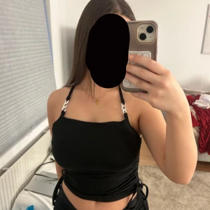 Svart linnetopp med kedjedetaljer från SHEIN - Svart linnetopp från SHEIN med kedjedetaljer på axelbanden och snörning i sidorna. Toppen är croppad och har en rak halsringning. Perfekt för dig som gillar edgy och trendiga plagg. Materialet är mjukt och stretchigt, vilket gör den bekväm att bära. Den är i storlek M men passar som S