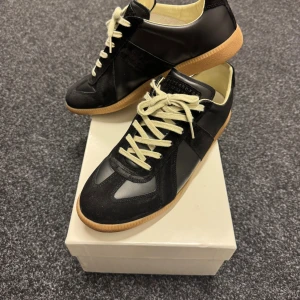 Maison Margiela GATS sneakers svarta - Maison Margiela Gats i svart med kontrasterande vita snören och beige insida. Snygga med rund tå och platt gummisula. Ovandel i skinn och mocka för en lyxig känsla. Kommer med originalkartong och dustbags.