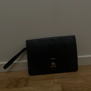 Svart kuvertväska i skinn med gulddetalj - Stilren svart kuvertväska i slätt skinn med guldfärgat lås framtill. Väskan har ett avtagbart handledsband och enkel, rektangulär form. Perfekt för att bära det viktigaste och ger en clean look till din outfit.