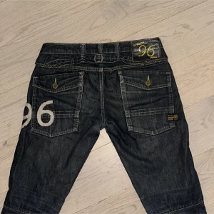 G-Star RAW mörkblå jeans med tryck - Snygga mörkblå jeans från G-Star RAW med coolt '96'-tryck på bakfickan och detaljerade sömmar vid knäna. Modellen har raka ben, stora bakfickor med knapp och klassisk femficksdesign. Materialet är slitstarkt jeans och passformen är normal.