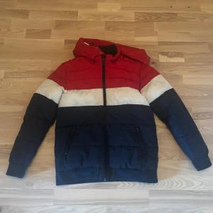 Färgglad pufferjacka från Levi's - Snygg pufferjacka från Levi's med färgblock i rött, vitt och mörkblått. Jackan har huva, dragkedja framtill och två fickor med dragkedja. Stor Levi's-logga på ryggen. Perfekt för kalla dagar när du vill sticka ut.