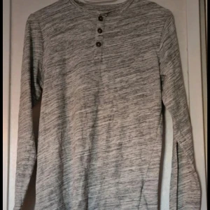 Grå långärmad tröja med knappar - Snygg gråmelerad långärmad tröja med tre knappar vid halsen. Tröjan har en enkel och stilren design med rund hals och ett diskret mönster. Perfekt för dig som gillar en chill och clean look.