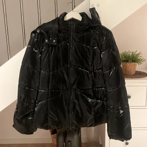 Svart pufferjacka från Guess - Säljer en glansig svart pufferjacka från Guess med huva och dragkedja framtill. Jackan har coola detaljer som Guess-logga på ärmen och metallmärke vid axeln. Perfekt för kalla dagar och riktigt snygg till streetwear-stilen.