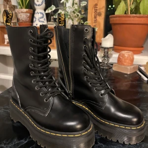 Svarta Dr. Martens boots med platåsula - Säljer ett par svarta Dr. Martens boots i skinn med grov platåsula och klassiska gula sömmar. Skorna har snörning framtill, dragkedja på sidan och rund tå. Perfekta för dig som vill ha en edgy och cool stil.