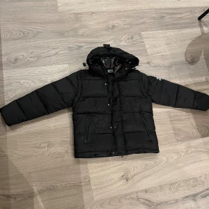 Svart pufferjacka från Burberry - Säljer en svart pufferjacka från Burberry med huva och dragkedja framtill. Jackan har två fickor med dragkedja och en snygg Burberry-logga på ärmen. Insidan har ett klassiskt rutigt foder i grått, vitt och rött. Perfekt för kalla dagar när du vill vara både varm och stilren.