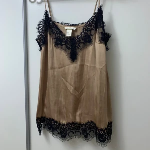 Beige linnetopp med svart spets - Säljer en beige linnetopp från H&M med tunna axelband och vacker svart spetsdetalj längs ringningen och nederkanten. Toppen har en mjuk, glansig yta och en avslappnad passform som känns både trendig och stilren.