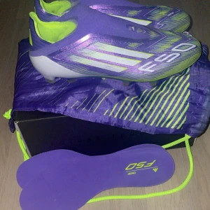 Lila Adidas F50 fotbollsskor - Säljer ett par lila och neongula Adidas F50 fotbollsskor med vita detaljer. Skorna har en aerodynamisk form, platt sula med dobbar och är tillverkade i syntetmaterial. Perfekta för dig som vill sticka ut på planen! Innersulor och påse medföljer. 