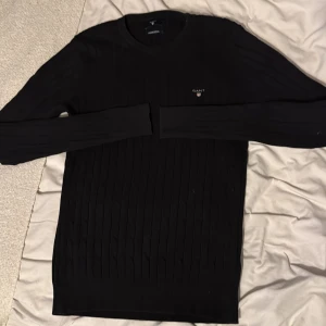 Svart kabelstickad tröja från GANT - Svart kabelstickad tröja från GANT i bomull med rund halsringning och diskret broderad logga på bröstet. Tröjan har lång ärm och ribbade muddar för en clean look. Perfekt för dig som gillar klassisk och stilren stil. Storlek M men sitter mer som S