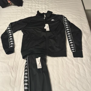 Svart Kappa ziptröja med loggor - Svart ziptröja från Kappa med vita loggor längs ärmarna och liten logga på bröstet. Tröjan har hög krage, dragkedja framtill och ribbade muddar. Tillverkad i glansig polyester för en sportig vibe. Perfekt för dig som gillar streetwear och sportiga outfits.