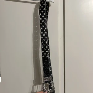 2st McBlimg bälten - Säljer ett brett bälte med glittrig yta i svart och silver, dekorerat med silvriga nitar och hål längs hela bandet. Spännet är i silverfärgad metall med detaljerat blommönster och westernstil. Perfekt accessoar för att lyfta vilken outfit som helst.