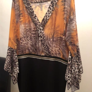 Mönstrad omlottklänning med leoparddetaljer - Säljer en unik omlottklänning från Coco Loco med coolt mönster i orange, grått och svart. Klänningen har leoparddetaljer vid halsringning och ärmar, samt breda, fladdriga ärmar. Nedre delen är svart med dekorativa ränder. Perfekt för dig som gillar att sticka ut!