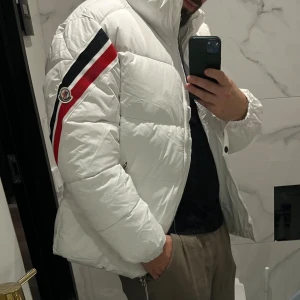 Vit pufferjacka från Moncler - Säljer en clean vit pufferjacka från Moncler med feta röda och svarta ränder på ärmen och klassisk logga. Jackan har dragkedja, hög krage och är riktigt varm. Perfekt för dig som vill sticka ut i vinterkylan.